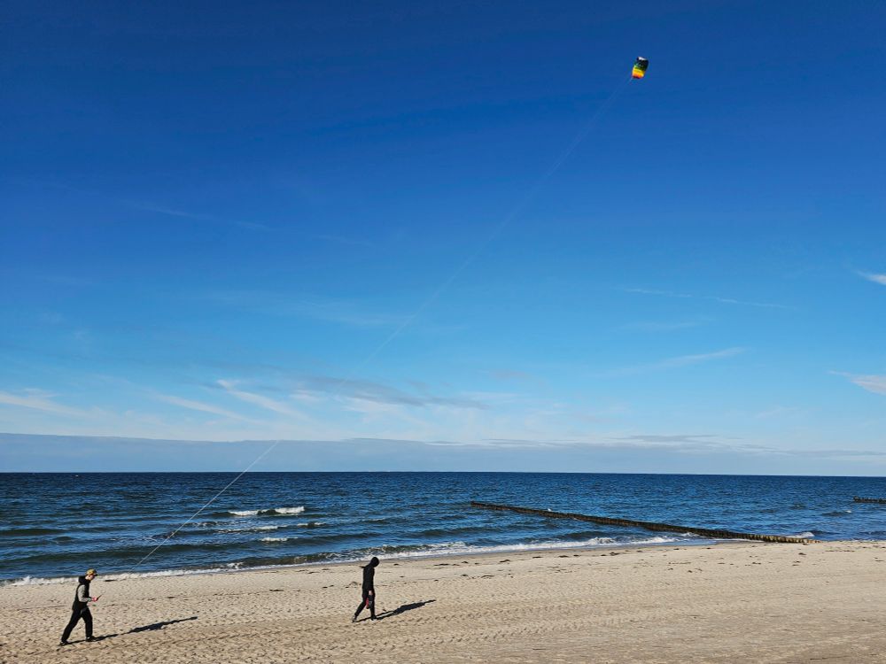 Das Bild zeigt eine ruhige Strandszene mit zwei Personen. Eine Person läuft am sandigen Ufer entlang, während die andere einen Drachen steigen lässt. Der weite blaue Ozean trifft am Horizont auf den klaren Himmel, über den helle Wolken verstreut sind. 