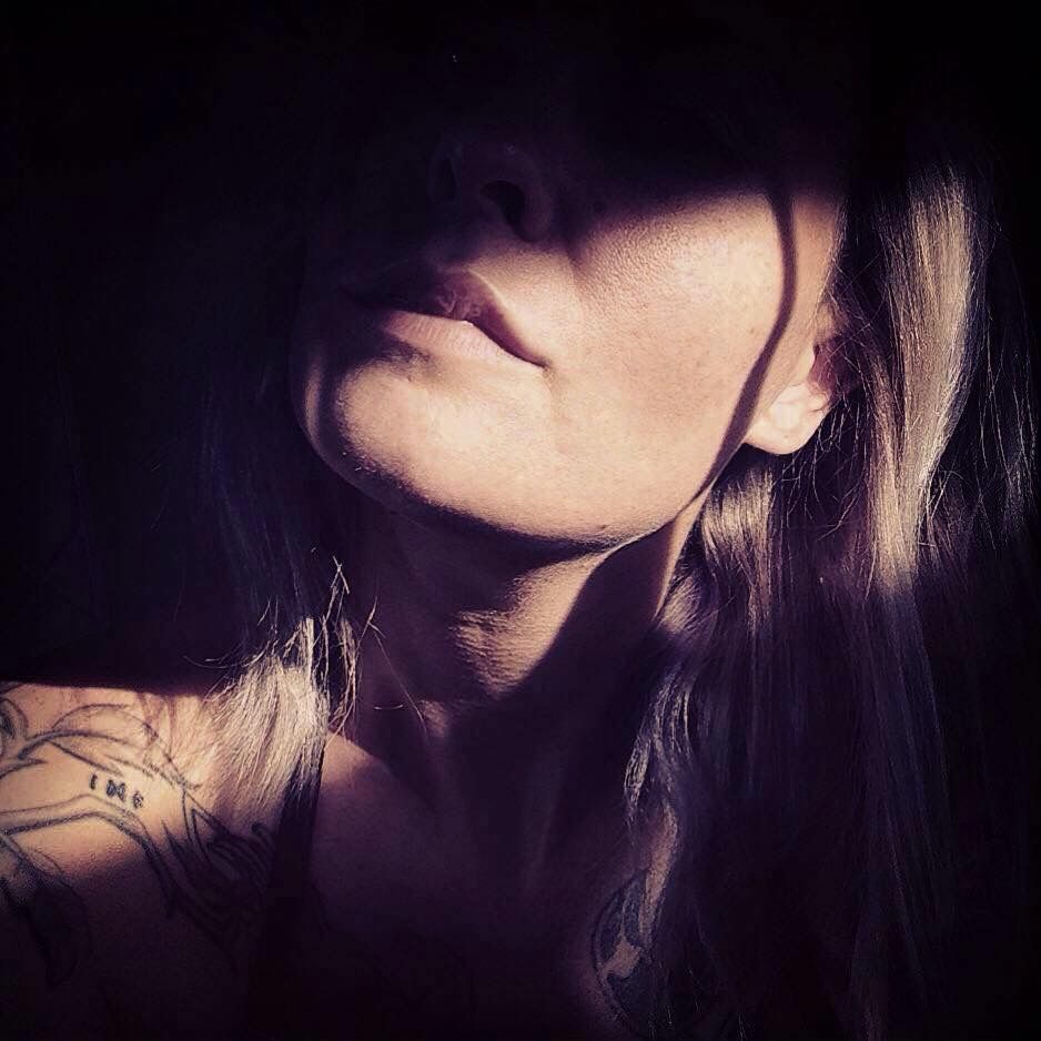 Nahaufnahme einer Frau mit langen, hellen Haaren und Tätowierungen auf der Schulter, teilweise im Schatten mit Licht, das ihr Gesicht teilweise beleuchtet.
