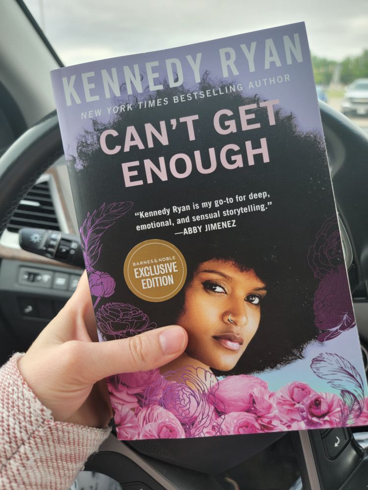 My hand holding the book Can't Get Enough by Kennedy Ryan which features a headshot if a gorgeous Black Woman with a giant afro surrounded by pink flowers on the cover