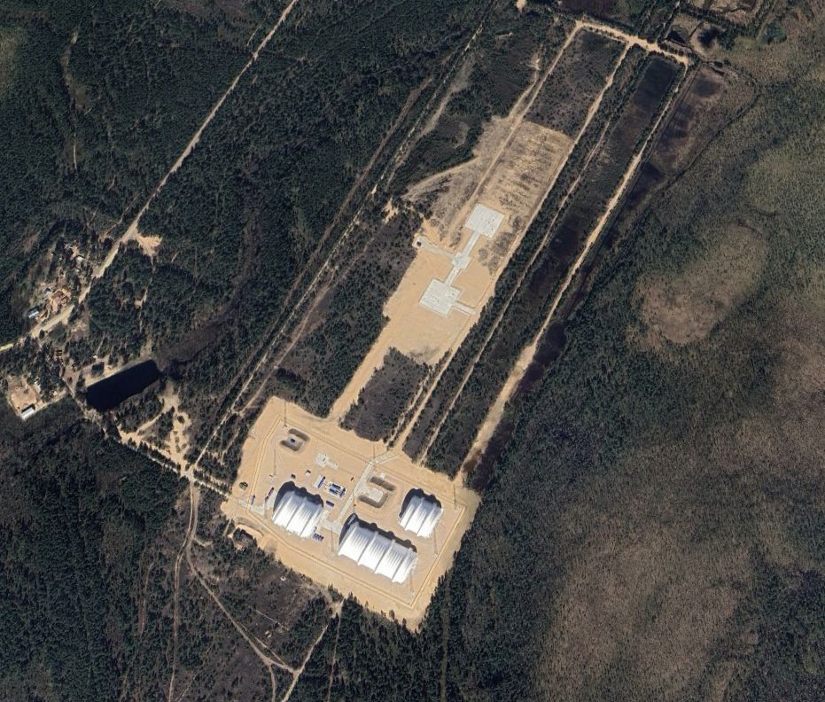 Site with lightning arrestor towers, twin layer fencing, large dome structures, one container surrounded on three sides by a berm, and four containers linked by piping in a central position. In addition to the north are two large concrete pads.