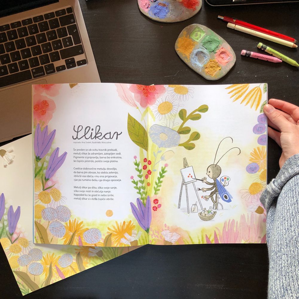 Photo from a bird perspective of a magazine spread lying flat on a black desk. There is an open laptop on the top left corner and art supplies on the right. A hand is holding the magazine.