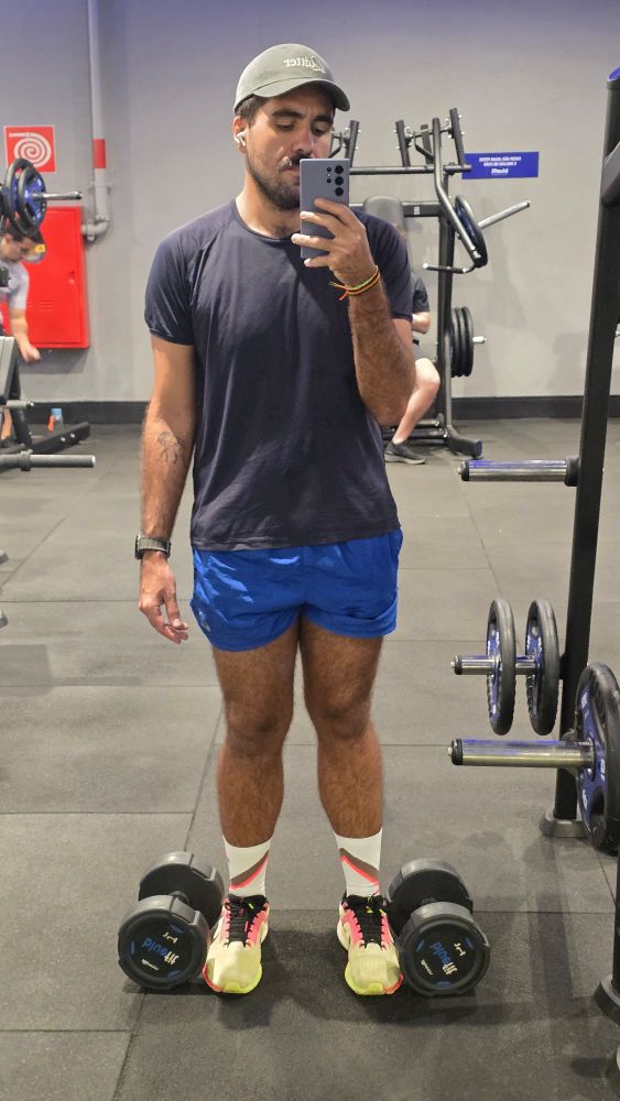Gay tirando selfie na academia com um shortíssimoazul, um tênis verde/rosa neon, camisa marinho e um bone cinza