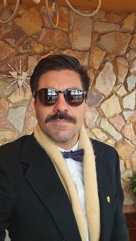 Gay de terno e gravata borboleta e óculos escuros 
No fundo uma parede de pedra e um divino espírito santo de madeira 