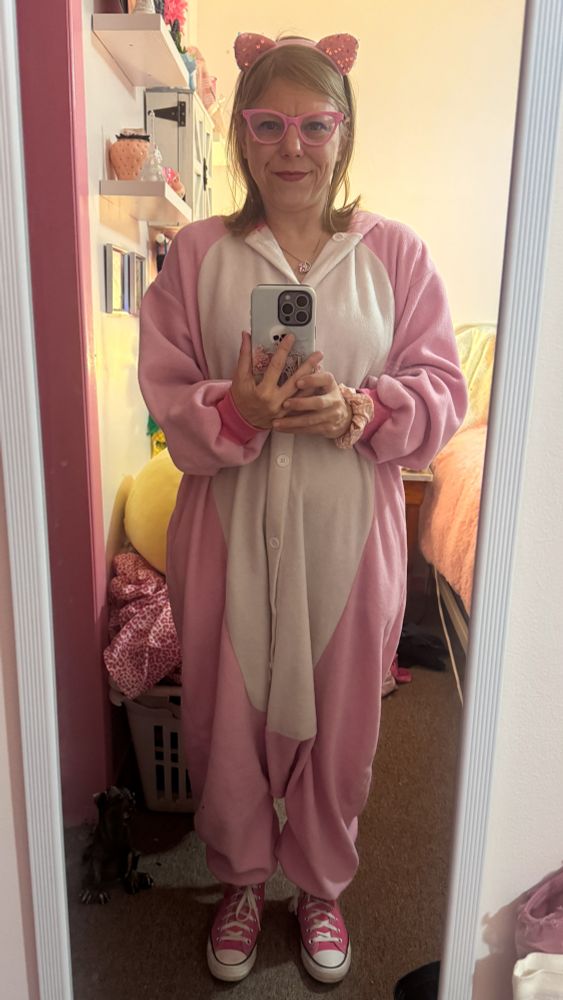 Sunny takes a mirror selfie in her bedroom while dressed in a cozy pink-and-white unicorn onesie. She’s wearing sparkly pink cat-ear headbands and pink cat-eye glasses, and she has on matching pink Converse sneakers. She holds her phone with a skeleton-themed case and smiles gently. The room around her has her usual pink decor, with shelves on the left and a bed with a fluffy pink blanket on the right.