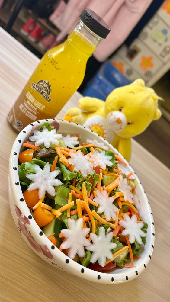 A photo of a white and pink ceramic bowl with black dots around the edges, full of salad with green leaf lettuce, snow peas; orange, yellow and red cherry tomatoes; cucumbers, matchstick carrots, shredded old cheddar cheese, and radishes cut into starburst shapes decorating the top. Beside the salad is a clear bottle of yellow lemon garlic vinaigrette, and a small, yellow, plush Funshine Care Bear.