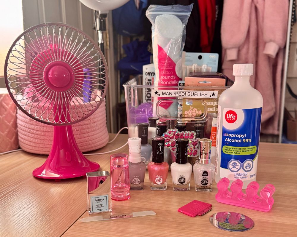 A photograph of a home manicure/pedicure setup sitting on a desk made of light wood. On the left side of the picture, you can see 1/4 of a light pink lamp with a white lampshade and a pink woven basket. In front of the basket, there is a small oscillating fan with a hot pink base and fan blade. To the right of the fan, there is a transparent plastic bin with a black and white label that says "MANI/PEDI SUPPLIES" in capital letters, containing nail polish remover, cotton rounds, bottles of nail polish and other assorted mani/pedi sundries. In front of the bin are four nail polish type bottles of Sally Hansen Miracle Gel products: primer, pink Step 1, white Step 1, and a glitter Step 2 top coat. Next to the top coat is a large bottle of 99% Life brand isopropyl alcohol. Directly in front of the alcohol is a pair of pink jelly toe separators, metal nail art stamp plate and scraper. To the left of those, on the other side of the Miracle Gel bottles, is a transparent pink nail art stamper, a tiny square bottle of Butter London brand Melt Away Cuticle Exfoliator, and a glass Butter London brand cuticle pusher thing.