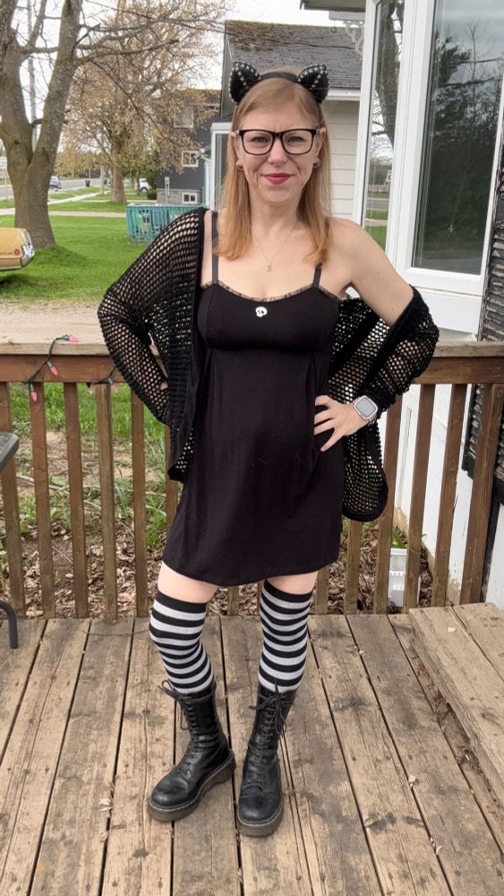 Sunny stands on a wooden porch wearing the black “Maila” nightie by Inspiration Vintage, layered with a loose black mesh cardigan. She has on black-and-grey striped thigh-high socks, black calf-high Doc Martens, and a black cat-ear headband. She’s smiling confidently with one hand on her hip, wearing black-framed glasses and a smartwatch. The background shows trees, houses, and a parked vintage car.
