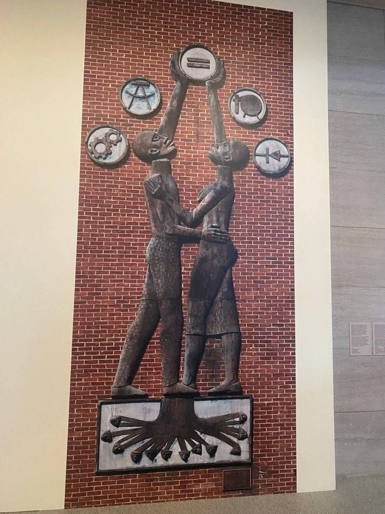 The Howard u engineering building sculpture by Elizabeth Catlett. A man and woman embrace while holding equal sign surrounded by other engineering tools while they stand on tree root stemming from African masks standing in as ancestors 