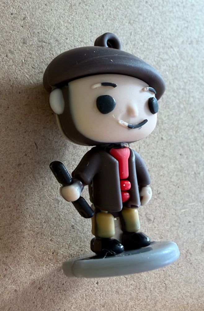A Funko Pop style figure with a flat cap and a chinstrap beard, a red shirt under a brown jacket, holding a stick.