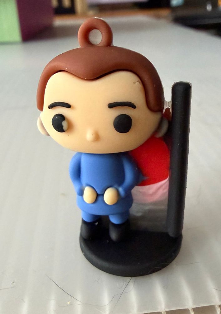 A Funko Pop style figurine wearing a solid blue outfit and standing next to what might be a red flag or just a manufacturing issue. 