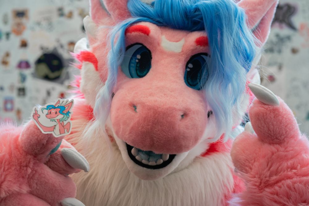 pink dragon fursuiter holding up a sticker of herself 'flipping the bird' while she also 'flips the bird'