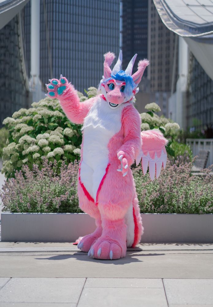 Pink dragon fursuit with blue hair standing in front of a flowerbed on the rooftop of the DLCC at Anthrocon 