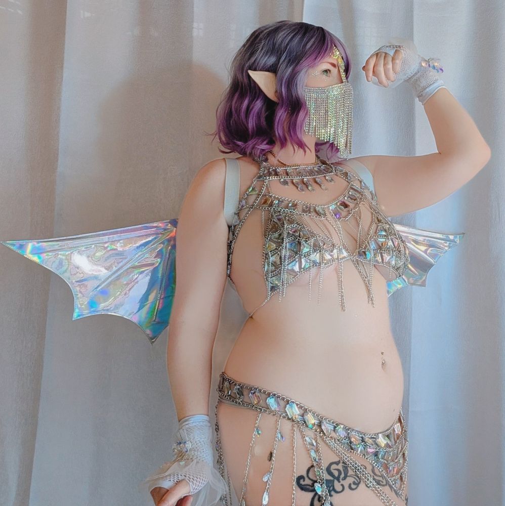 cosplaykinky side profile in a jeweled silver chain bikini ensemble, thigh garter with star details, holographic dragon wings, jewel veil and short wavy pink and purple hair.
