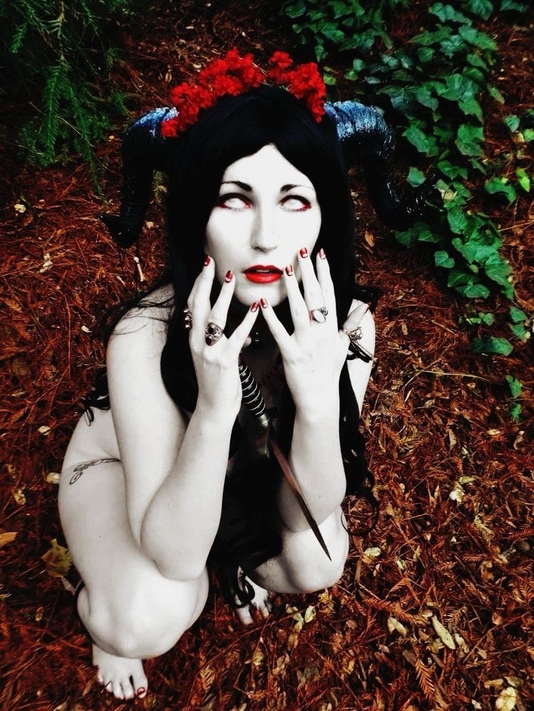 cosplaykinky cosplaying Aradia Megido as a ram god with black horns. they're crouching looking up at the viewer, naked but tastefully concealed by their hair, hands raised, wielding a blade.