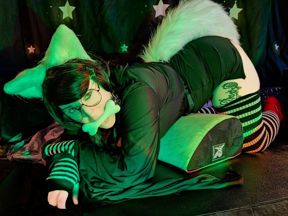 cosplaykinky as dog tier Jade Harley bent over a pillow on elbows and knees, staring heatedly at the viewer with a dog bone gag in their mouth.