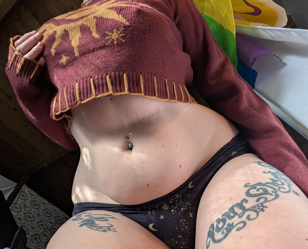 cosplaykinky looming over the viewer in a maroon long sleeve crop with a sun printed on the chest and purple undies with cosmic patterns. their hand is casually laid over their breast.