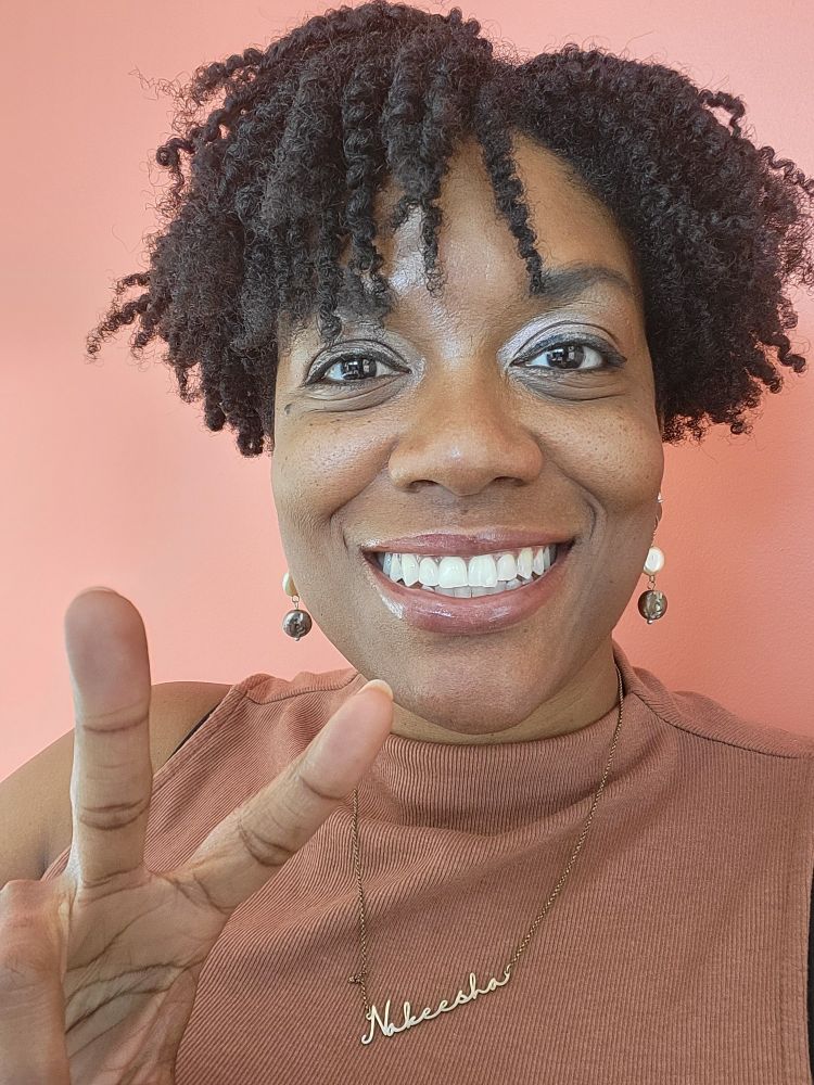Selfie of Black woman with curly short black hair
 Smiling, wearing a brown ribbed halter, white and silver earrings with a gold necklace spelling out "Nakeesha," holding up a peace sign with a deep peach colored wall as the background. 