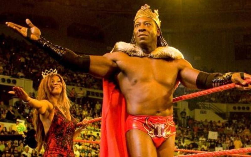 King Booker stands in a wrestling ring with Queen Charmel. He wears tights, a crown, a cape, and nothing else.