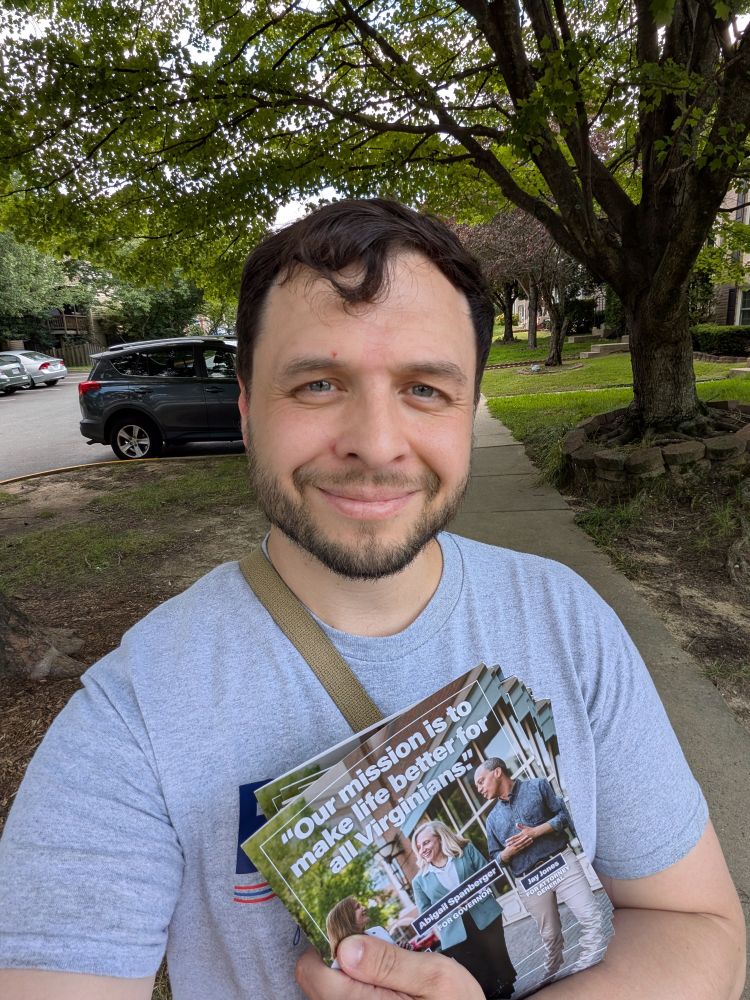 A man standing in a neighborhood with literature for the Virginia Statewide coordinated ticket. He is wearing a "Briana Sewell for Delegate" t-shirt.