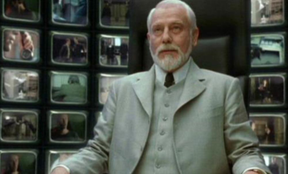 A man with white hair, white beard and a white suit, sitting on a tall black office chair, with many small screens lit up behind him. Scene from the matrix reloaded, the screens are playing other parts of the very movie
