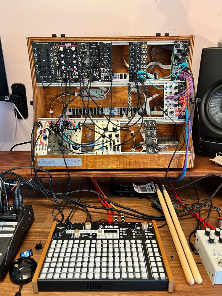 A photo of my hardware synthesizer setup, including a eurorack module case, a Synthstrom Deluge, a digital mixer, and many cables in between. It is spread out on a solid wood desk with a shelf at the back for the wooden module case