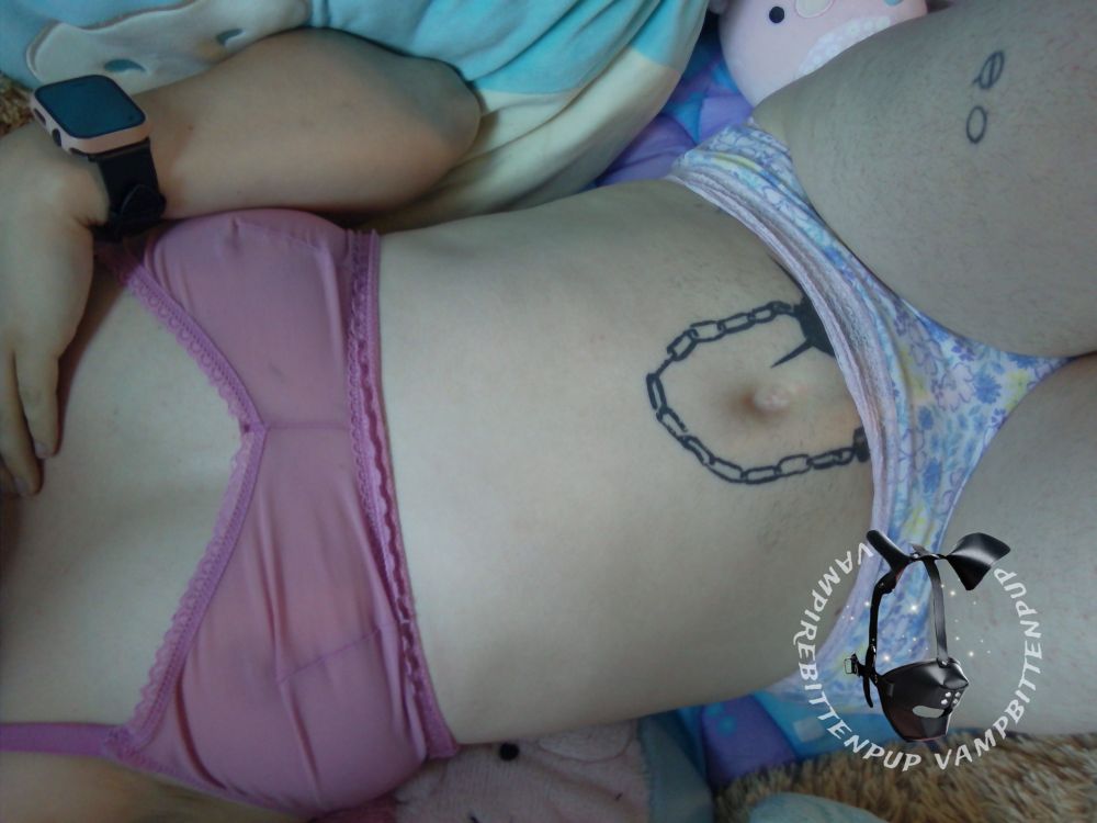 image of bingo bitten laying back against colorful baby blankets and large plush toys, one hand daintily rested on his collarbone, back arched forward and  furry thighs squished together. a large black mace tattoo pokes out above his pastel floral lace panties, and his pink nipples are visible through his sheer pink mesh and lace bralette 