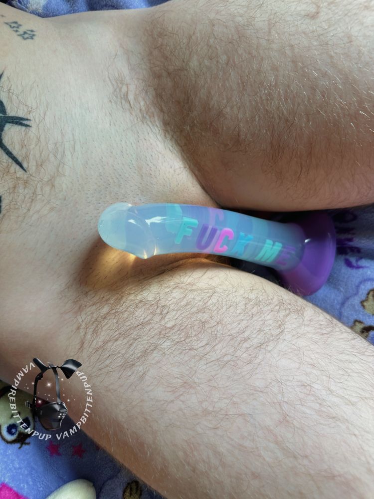 image of bingo’s mid thighs to lower tummy, as he’s laying back on his bed. he’s nude, with fur allll over except for his shaved cunt. a large black mace tattoo and small star cluster tattoo decorate his tummy. his soft thighs are spread, with a pretty dildo with the words „fuck me” on it sitting in between 