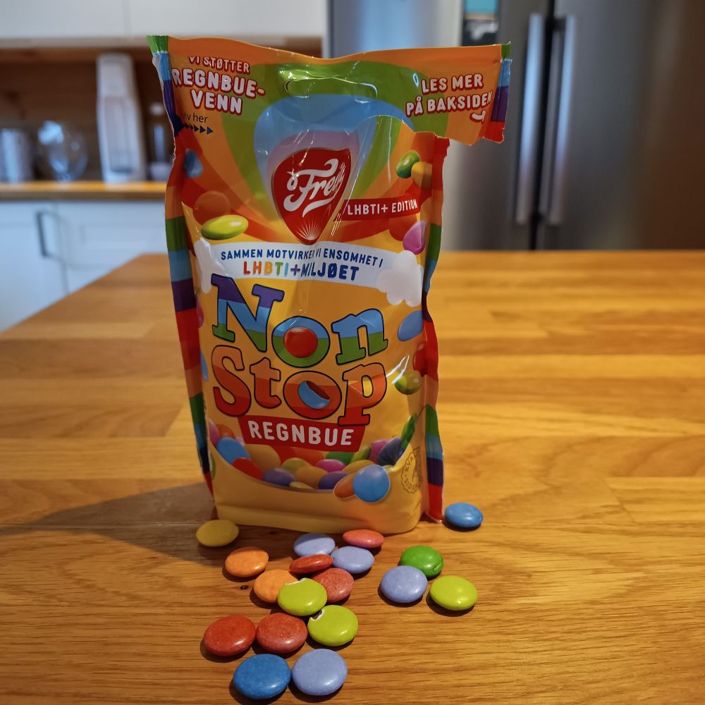 En åpen pose med regnbue-NonStop stående på en trebenk, med en liten håndfull NonStop spredt rundt pose.