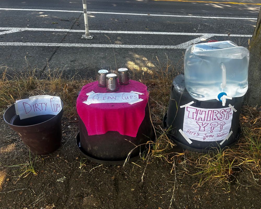 Photo of three small tables on the side of the sidewalk, one with the water jug and spout with the sign reading “thirsty?? There’s some water”, a second table with a sign reading “clean cups” with the accompanying cups, and to the side of that a bucket with a sign reading “dirty cups”