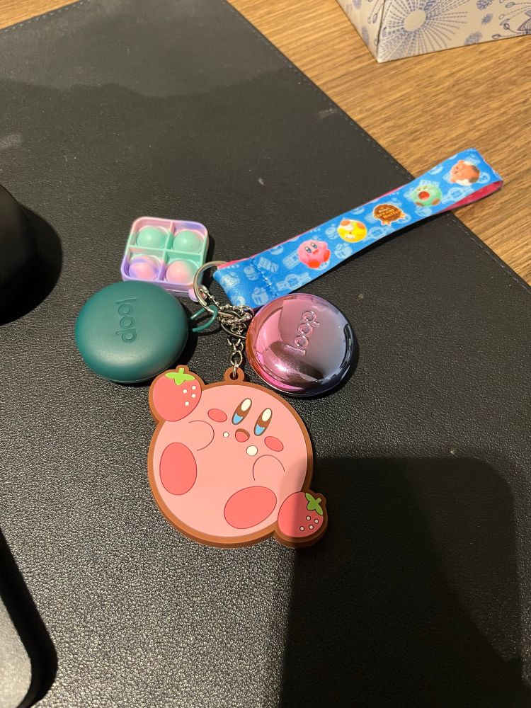 A keychain/lanyard with a rubber Kirby pendant, a tie dyed pop-it fidget, and two carrying cases for loop ear plugs