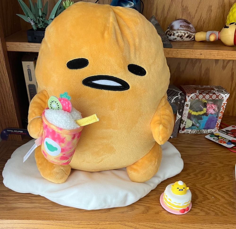 A Gudetama plush from build-a-bear workshop holding a smoothie, next to a tiny figure of Gudetama popping out of a cake 