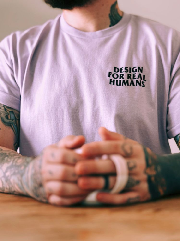 A light pink shirt says “design for real humans” on the top right. It’s modelled by Scott and his massively tatted hands and arms 