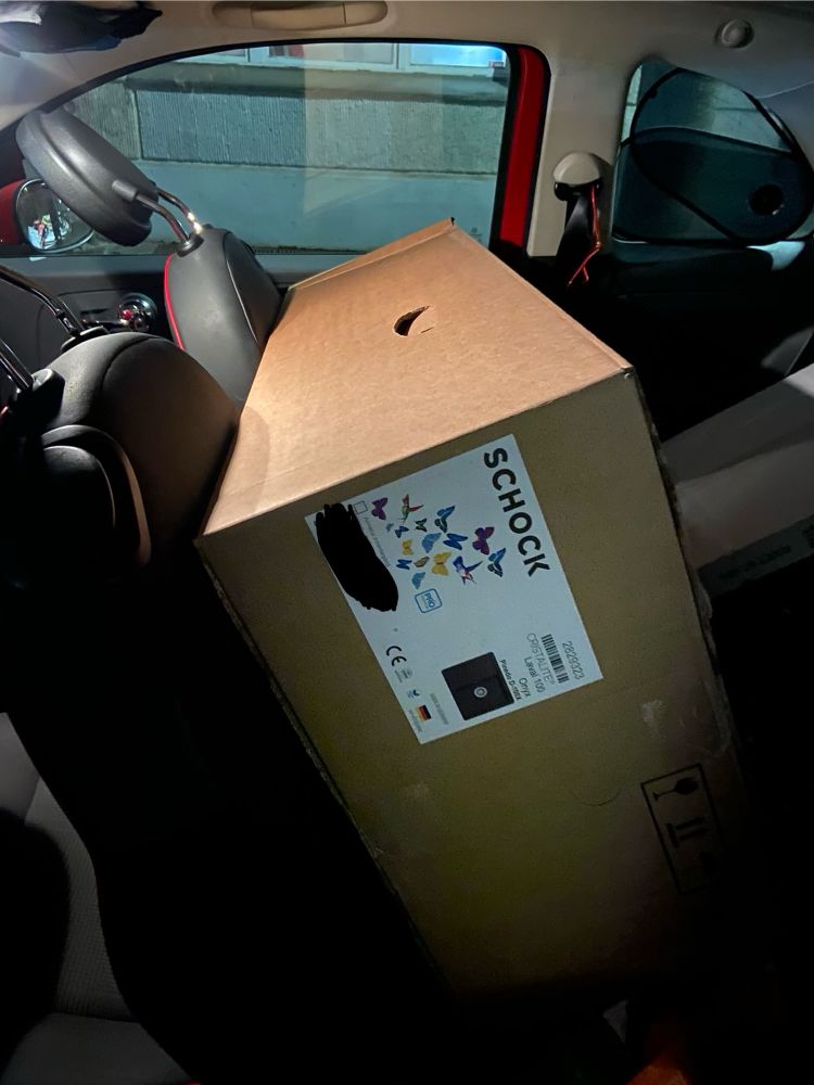 ein sehr großes paket mit einem aufkleber der firma „schock“ und schmetterlingen ist in ein kleines auto gequetscht. man sieht die beiden vordersitze umgeklappt und das paket dahinter ist fast so lang wie die rückbank breit