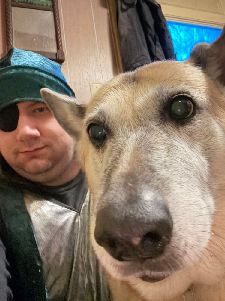 futureblues and Cara the german shepherd mix taking a selfie. futureblues is wearing a black cloak with a shiny silver stripe vertically down the front, a green and black cap with a very long top, and an eyepatch. Cara is close to the camera—looking forward.