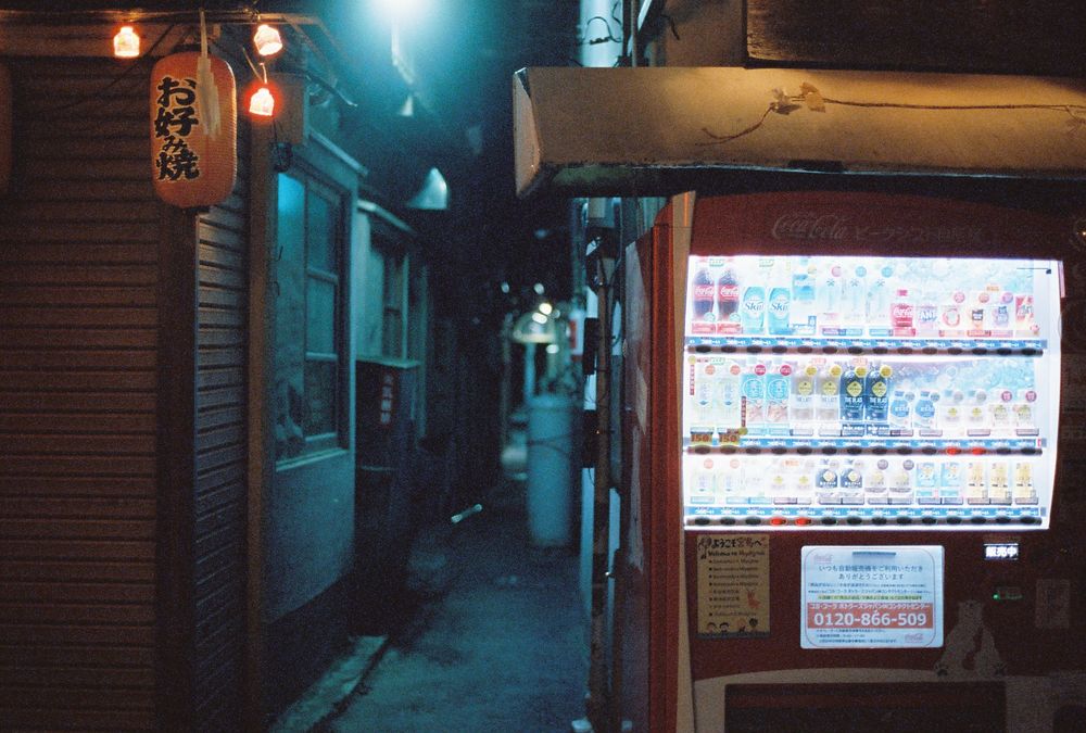 Bright vending machine next to narrow alleyway lit by blue lighting.