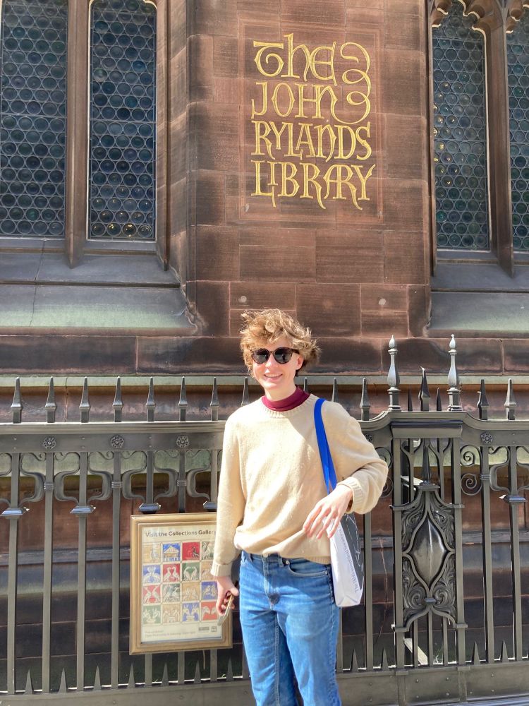 Finch, a white person with short brown hair, standing in front of the John Rylands Library, a red stone building with an ornate fence and the library’s name in gold on the side. Finch is smiling at the camera. 