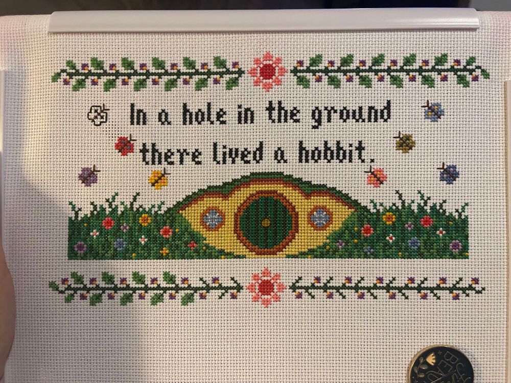 Cross stitch WIP on white aida cloth. Shows veins with green leaves and purple/yellow flowers, with a stylized red and pink flower in the center. Below is the text “in a hole in the ground there lived a hobbit.” Below that is a hobbit hole entrance with yellow wall, blue windows, and green door. Grass on the left and right sides with various pink, blue, purple, yellow, and white flowers. On the left are a purple butterfly, yellow butterfly, red butterfly, and white and black butterfly above the grass. On the right above the grass are a pink butterfly, blue butterfly, a golden butterfly, and a light blue butterfly. 