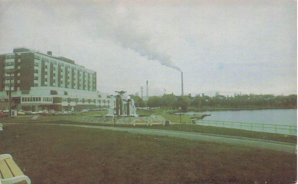 carte postale représentant un paysage glauque aux tons verdâtres, avec un banc partiellement visible en premier plan, une pelouse sombre, un plan d'eau, un bâtiment moderne et au loin une cheminée crachant de la fumée sur un ciel blanc. 