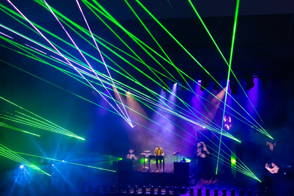 Masse grønne, hvite og blå lasere peker opp i luften over Jean-Michel Jarre som står på scenen, omringet av synthene sine.