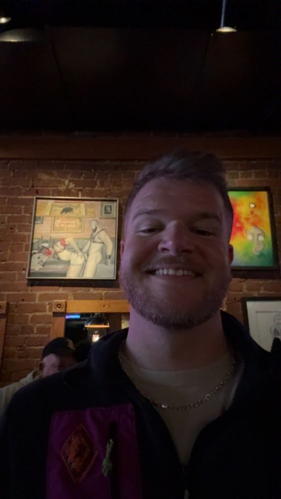 A photo of myself smiling, in the background is a painting of two men engaged in sex, on a brick wall