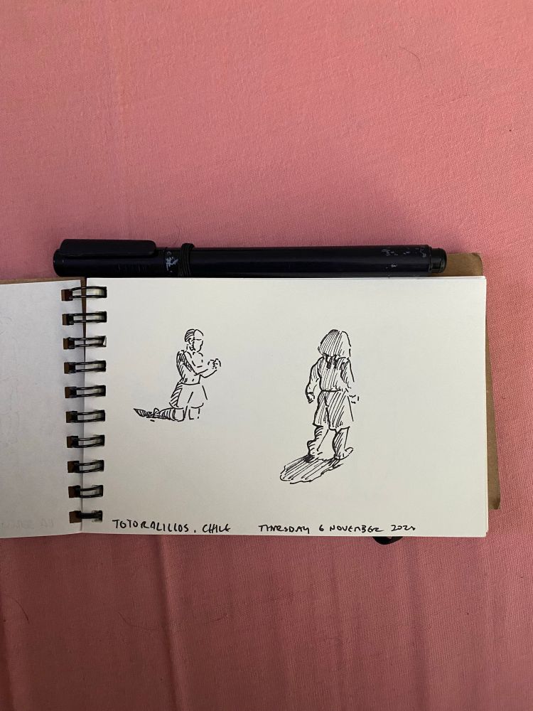 A photo of a notebook with two little figures drawn in ink pen. One is a boy playing in sand and another is a small child with long hair walking away from the artist. 