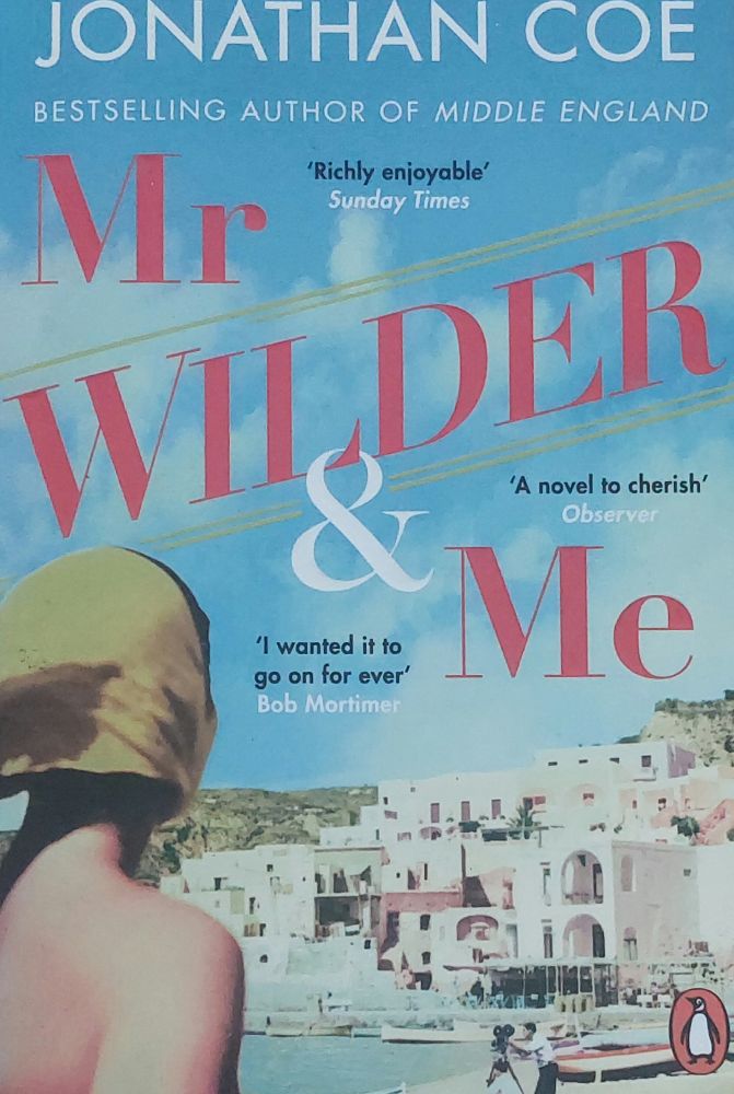 The bare shoulders and back of a young woman on the left. She is looking across the beach of a small Greek island with apartments in the background. On the beach a cameraman is filming. 