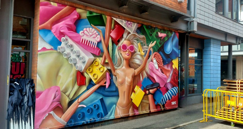 Brightly coloured mural featuring 'Barbie' doll with raised arms and wearing pink sunglasses. Background is a mix of Lego and Barbie accessories. 