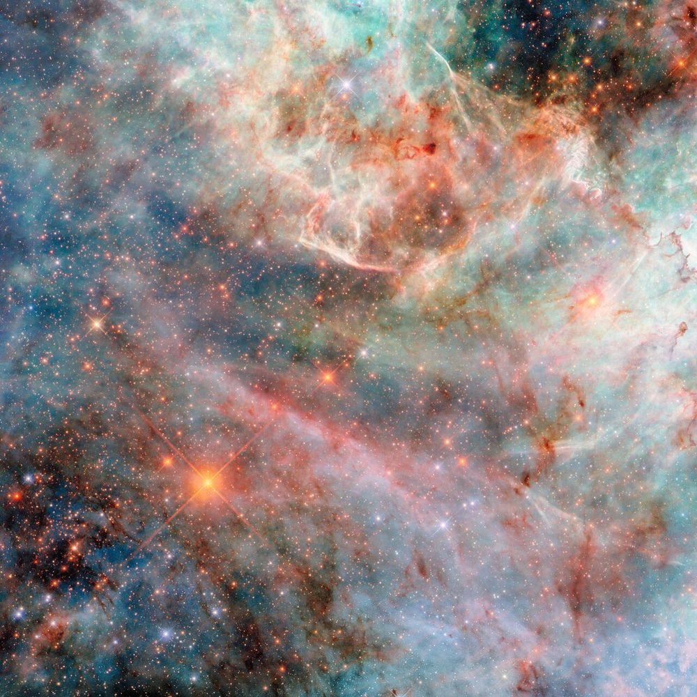 Description: A part of a nebula in space. It is made of layers of gas and dust clouds in different colours, from blue and green shades to pink, red and black, indicating light emitted by different molecules. The background cloud layers are thicker and puffier, though still translucent, and the upper layers are thin and bright at the edges. Behind the clouds are very many small, mostly orange and some blue, stars