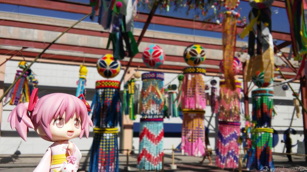 Nendoroid yukata version Madoka Kaname, standing to left of multiple Tanabata kazari decorations, as she glances right, in their direction. 