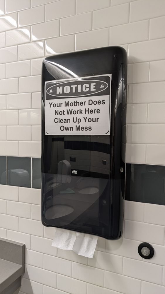 A printed sign placed on a paper towel dispenser in a men's restroom reads: Notice - Your Mother Does Not Work Here Clean Up Your Own Mess.