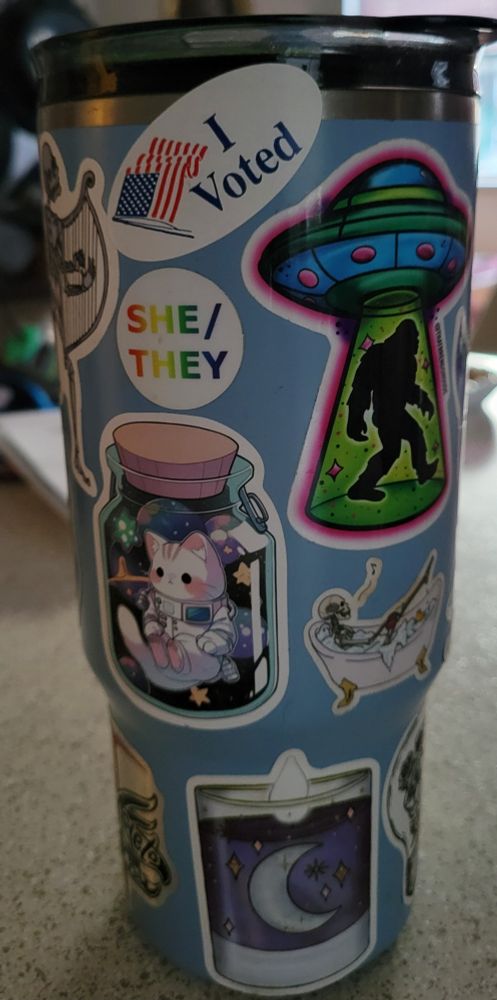 A blue coffee travel mug covered in stickers. "I voted" is at the top. Other notable stickers: an astronaut kitty, a skeleton in a bathtub, Bigfoot in a UFO's beam, she/they pronoun sticker. 