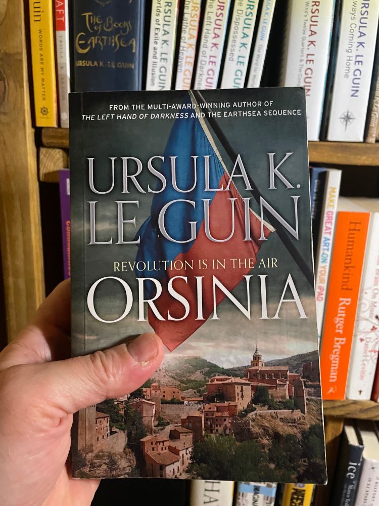 A while male hand holding up 'Orsinia' by Ursula K. Le Guin in front of a shelf of Le Guin's other books.
