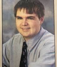 A yearbook photo of a much wider- sized boy with an ill-advised haircut, without a hint of curl. 

He’s wearing a light blue/gray shirt with a dark-colored tie. 
