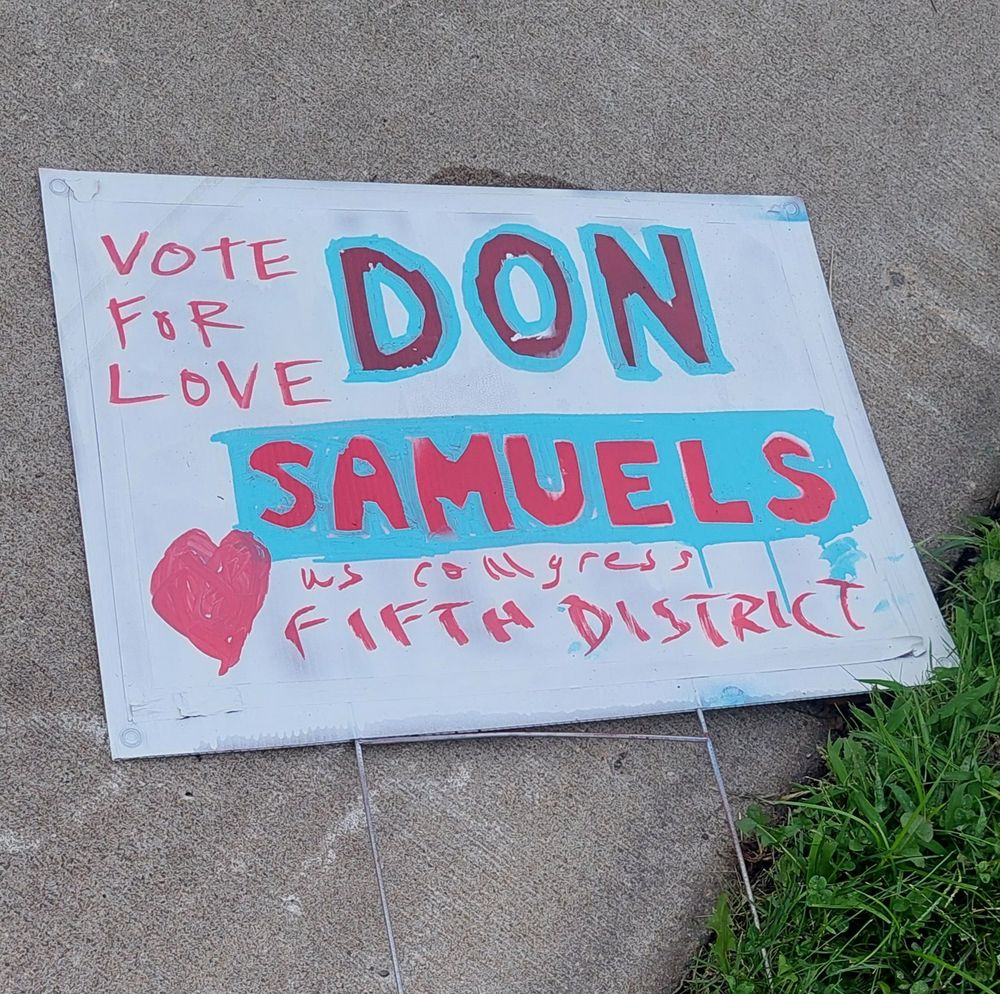 Handpainted yard sign lying on the sidewalk. The sign reads "Vote for Love" in the upper left corner. There is a red heart drawn in the lower left corner. The rest of the sign reads "Don Samuels" in large red and blue letters with "US Congress Fifth District" scrawled underneath it in red.